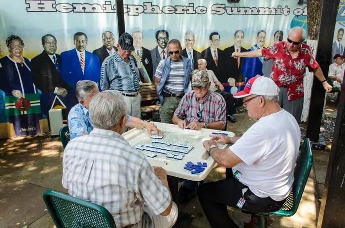 Grupo de hombres mayores jugando dominó en un parque, con mural de fondo.
