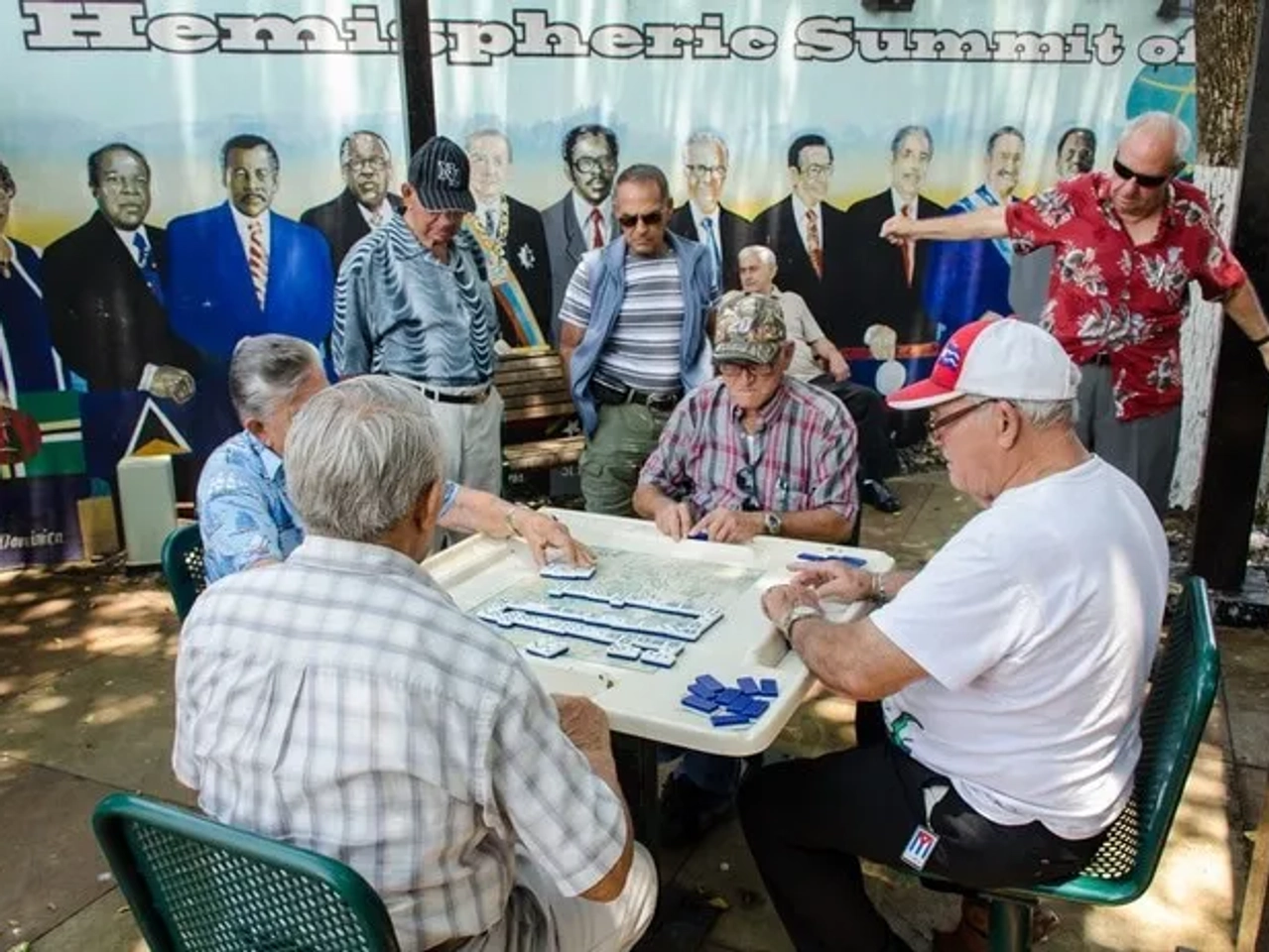 Grupo de hombres mayores jugando dominó en un parque, con mural de fondo.