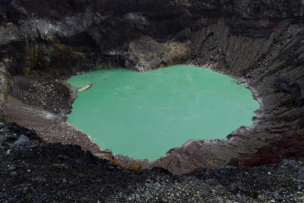 Cráter volcánico con agua verde en su interior, rodeado de rocas.
