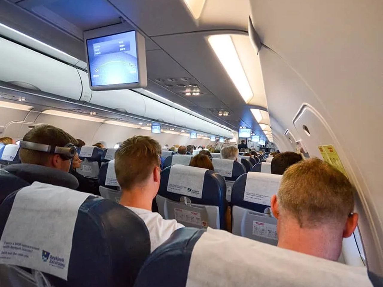 Pasajeros en un avión, con pantallas de entretenimiento y asientos ocupados.