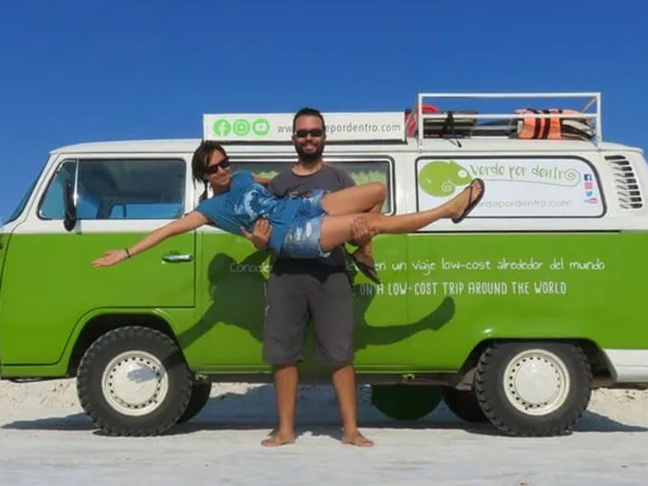 Pareja sonriente frente a una furgoneta verde en un paisaje desértico.