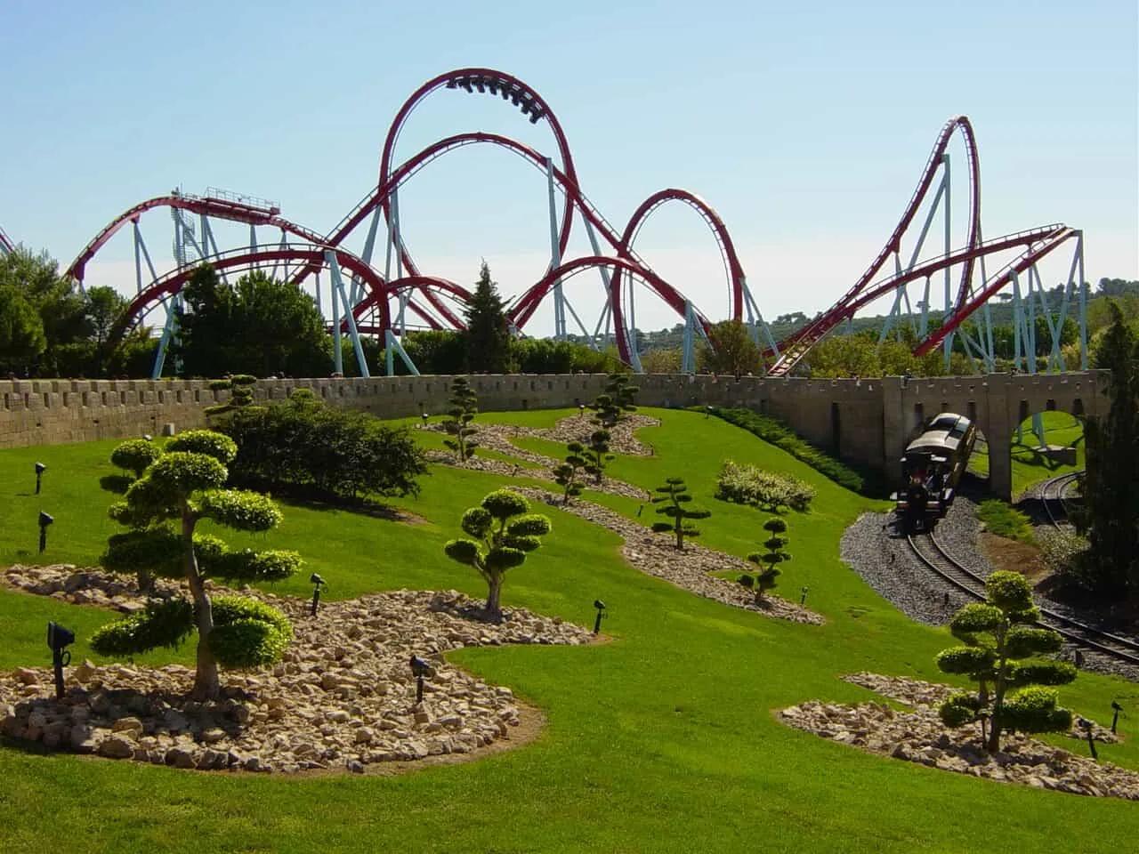 Montanha-russa imponente em parque de diversões, com paisagismo e trilhos de trem.