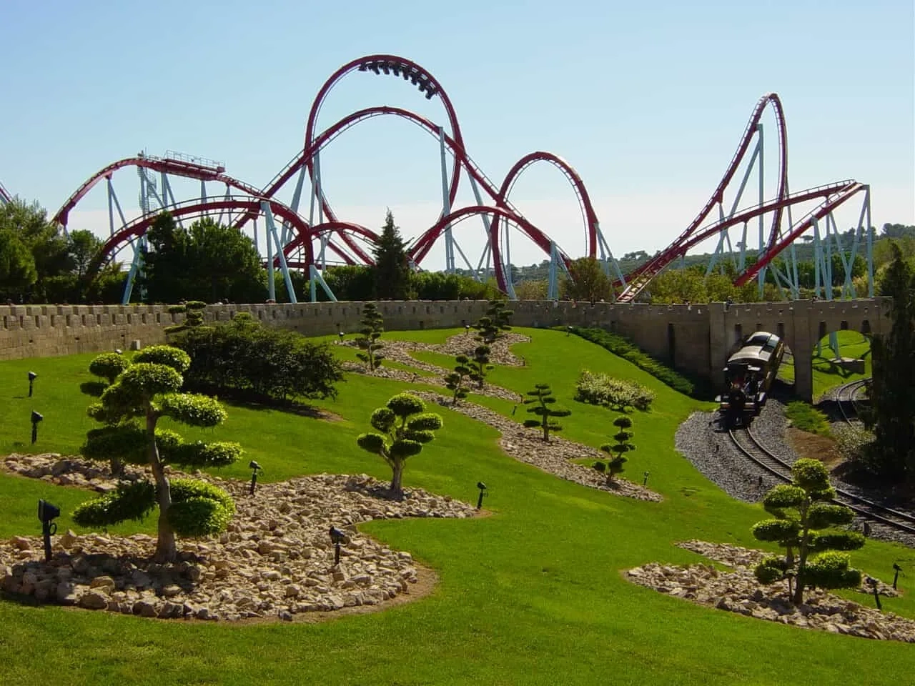 Montanha-russa imponente em parque de diversões, com paisagismo e trilhos de trem.