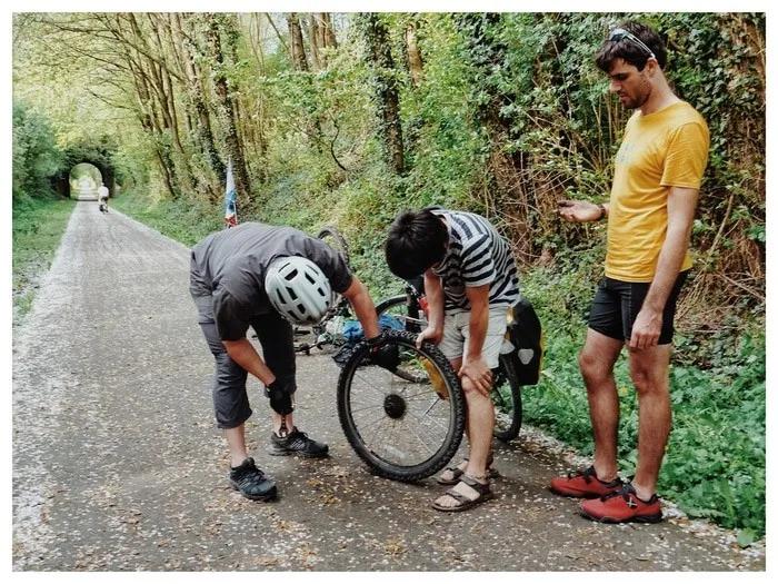 Tres personas reparan una bicicleta en un camino rodeado de árboles.