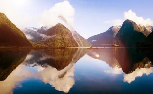 Paisaje montañoso con reflejos en el agua y un barco en la distancia.