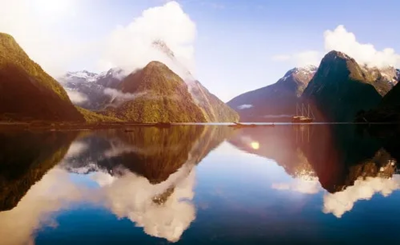 Paisaje montañoso con reflejos en el agua y un barco en la distancia.