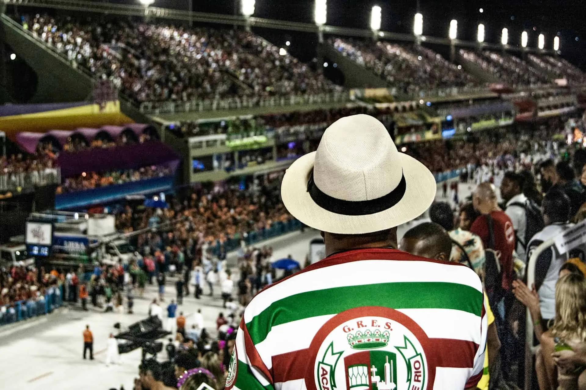 Homem com chapéu observa desfile de Carnaval em estádio lotado.