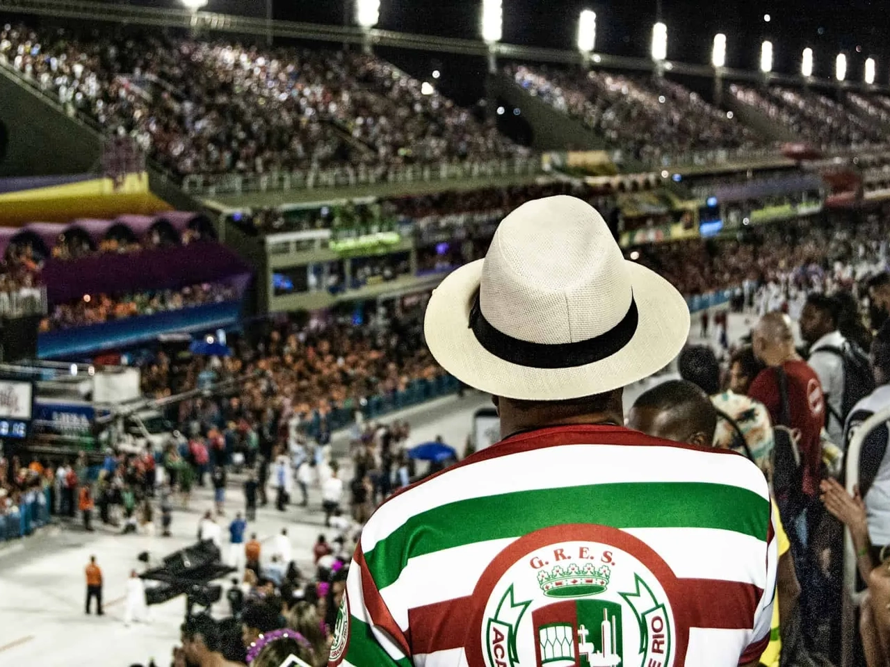 Homem com chapéu observa desfile de Carnaval em estádio lotado.