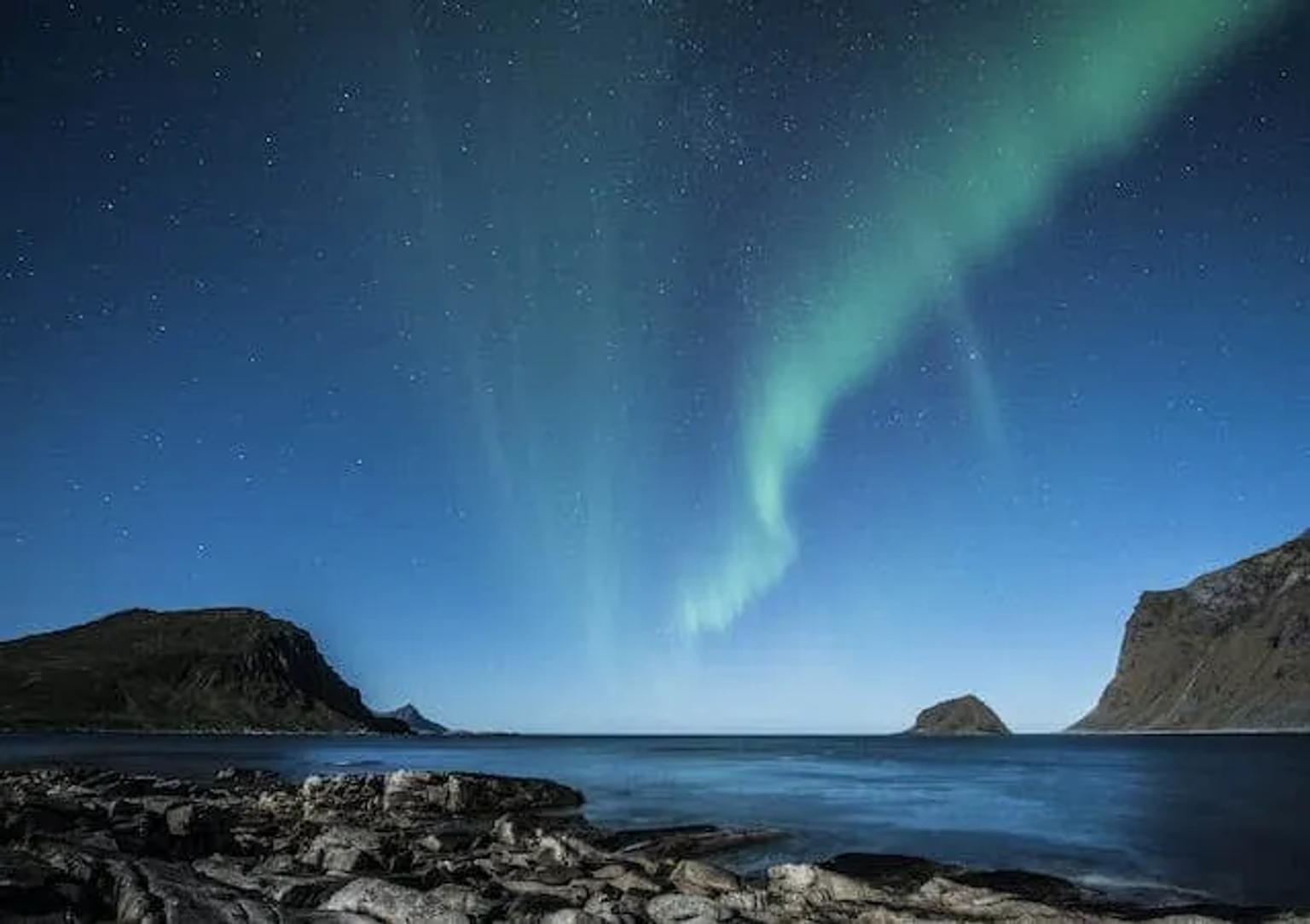 Northern lights illuminate the night sky over a rocky coastline and calm sea.