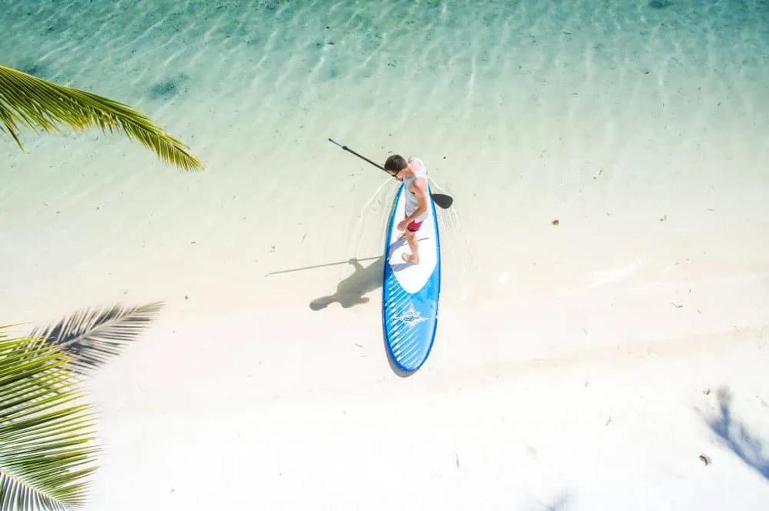 Persona practicando paddle surf en aguas cristalinas de Maldivas. Playa y palmeras al fondo.