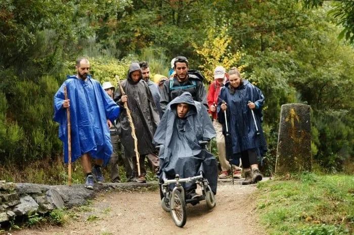 Grupo de personas con impermeables caminando por un sendero, acompañando a un joven en silla de ruedas.