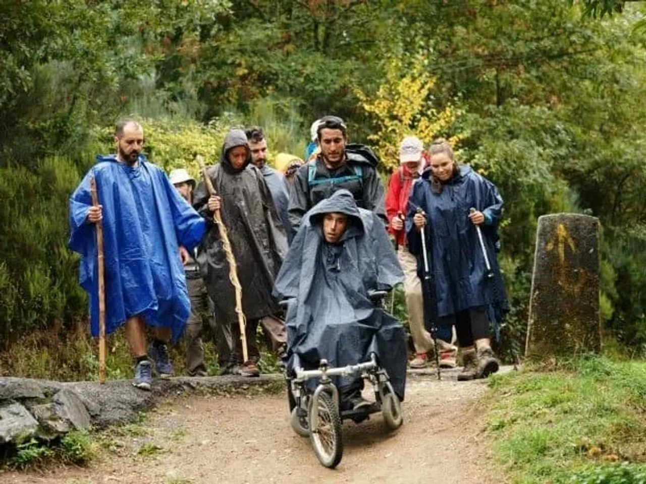 Grupo de personas con impermeables caminando por un sendero, acompañando a un joven en silla de ruedas.