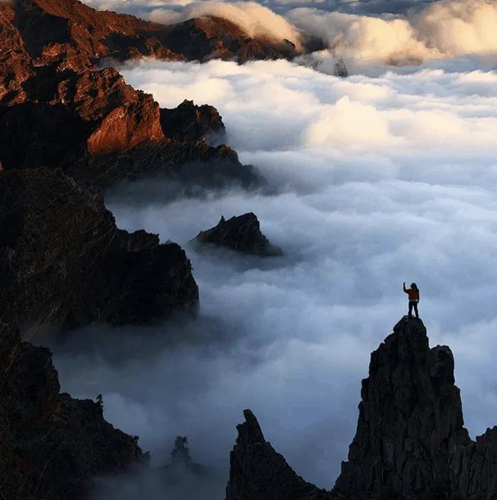 Persona de pie en una cima montañosa, rodeada de nubes y paisajes impresionantes.