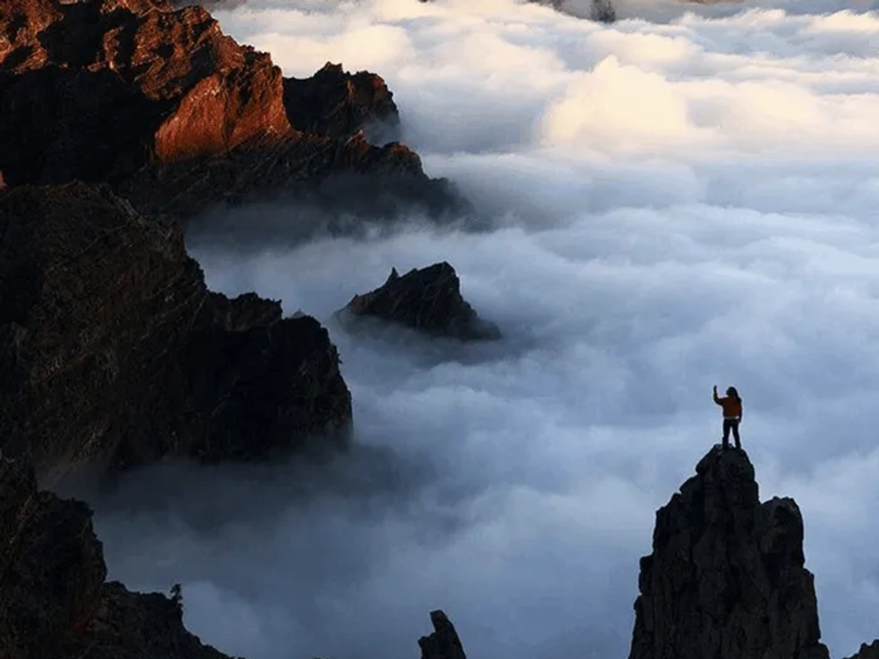 Persona de pie en una cima montañosa, rodeada de nubes y paisajes impresionantes.
