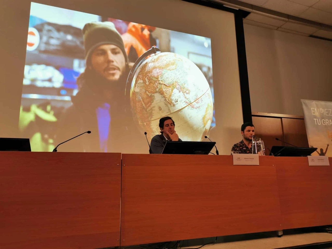 Dos ponentes discuten sobre comunicación y viajes, con un globo terráqueo de fondo.