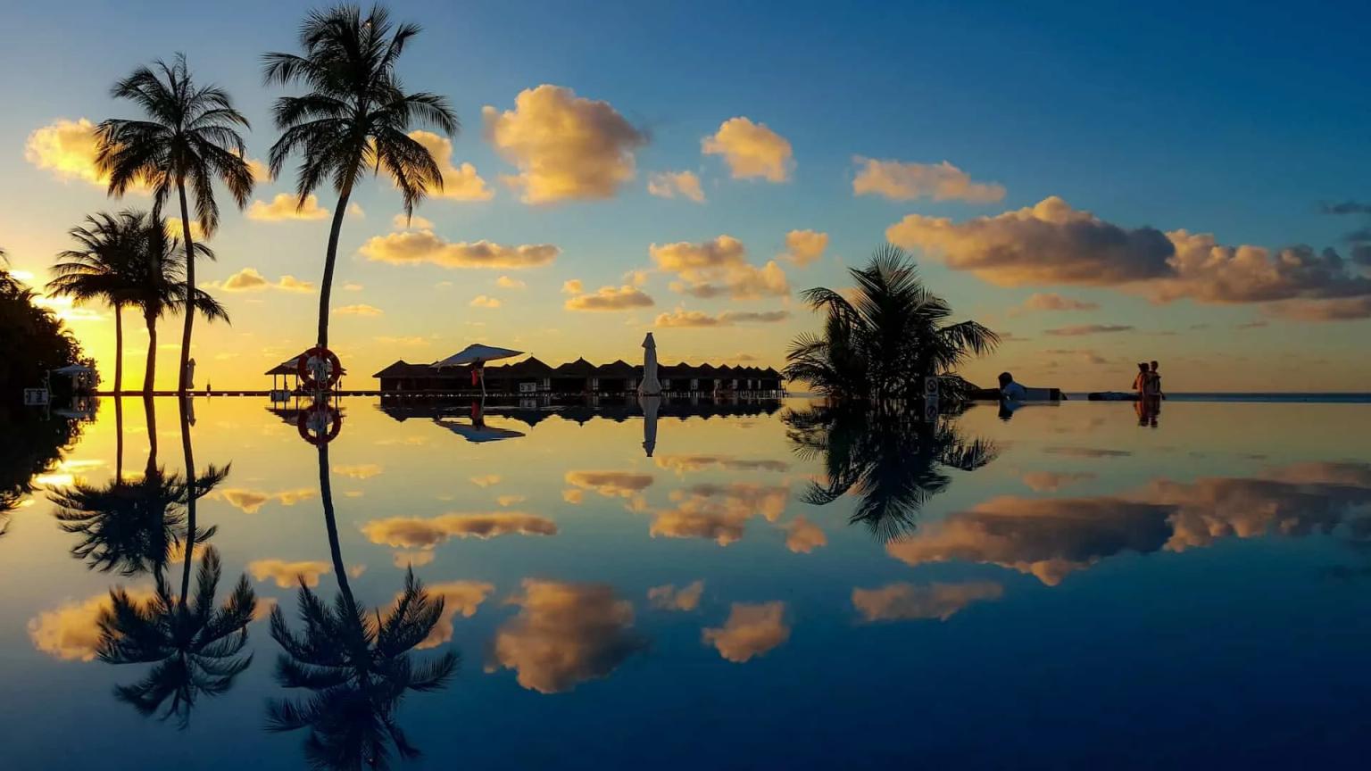 Pôr do sol refletido em piscina com palmeiras e bangalôs ao fundo.