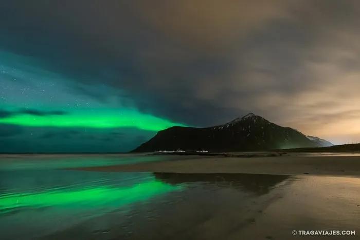 Auroras boreales iluminan el cielo sobre una playa y montañas reflejadas en el agua.