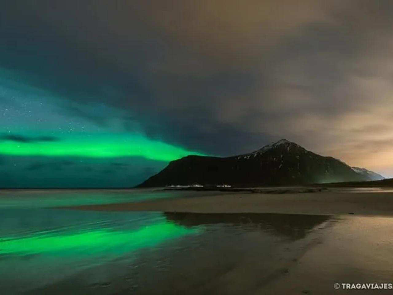 Auroras boreales iluminan el cielo sobre una playa y montañas reflejadas en el agua.