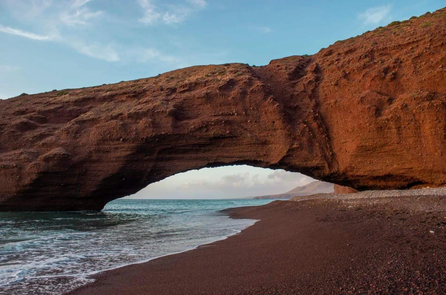 Arco natural de rocha vermelha na praia, com mar ao fundo.