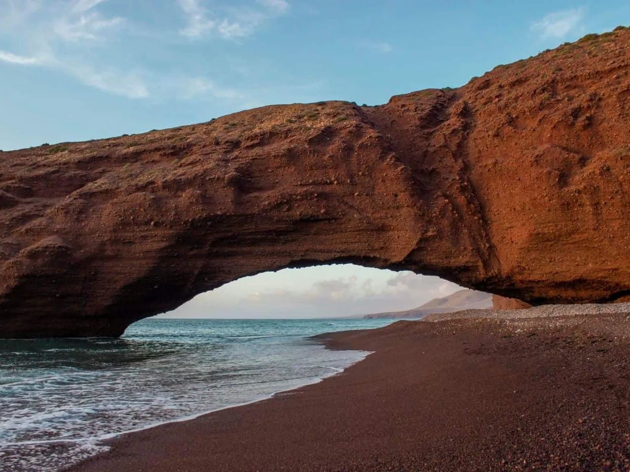 Arco natural de rocha vermelha na praia, com mar ao fundo.