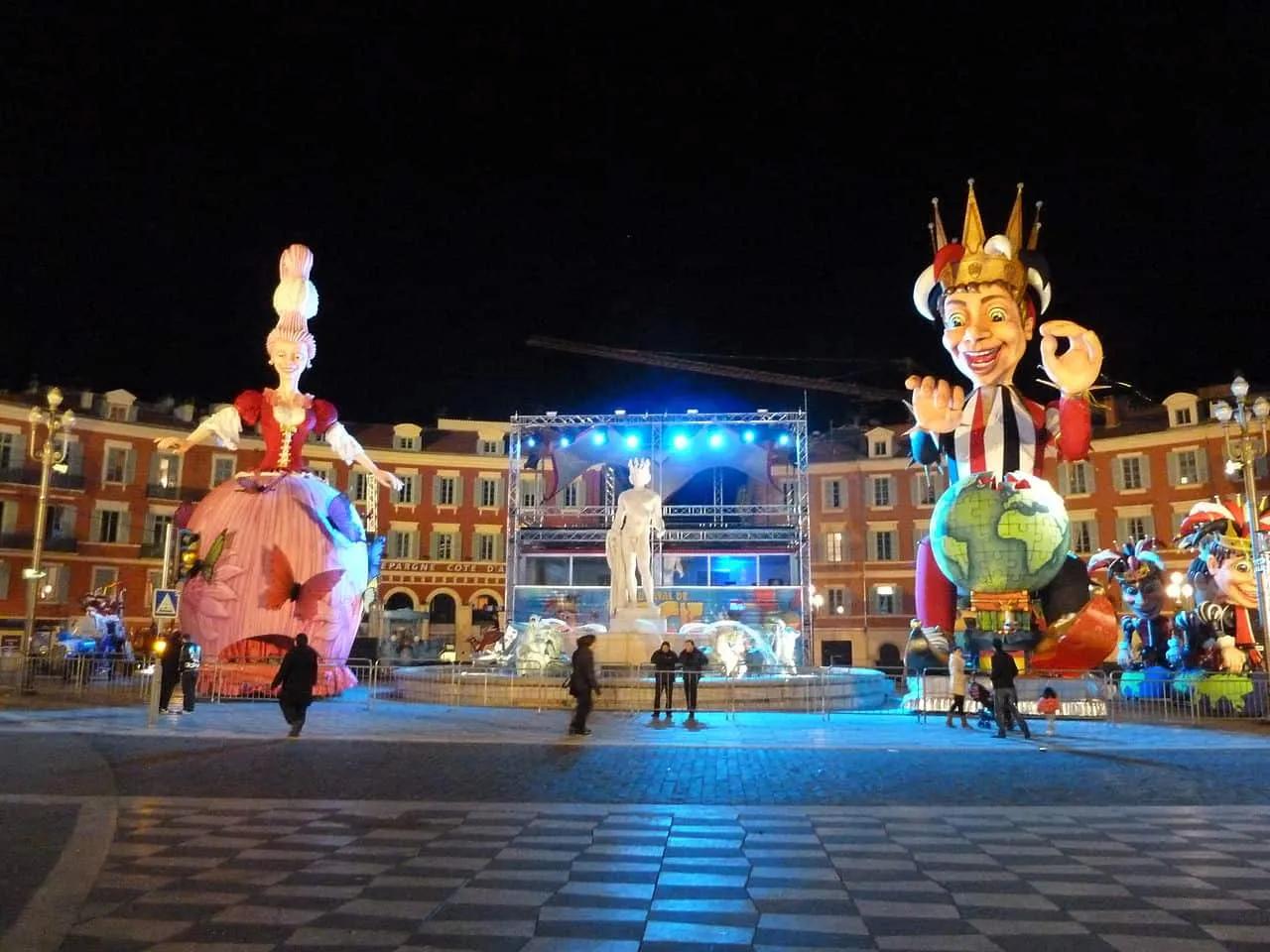 Desfile de Carnaval com grandes bonecos em uma praça iluminada à noite.