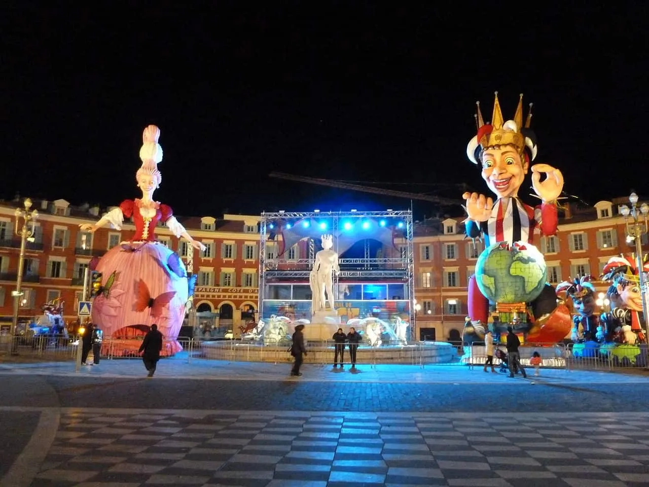 Desfile de Carnaval com grandes bonecos em uma praça iluminada à noite.