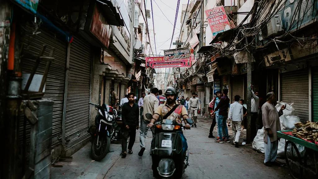 Rua movimentada em Nova Deli, com pessoas e motocicletas entre lojas fechadas.