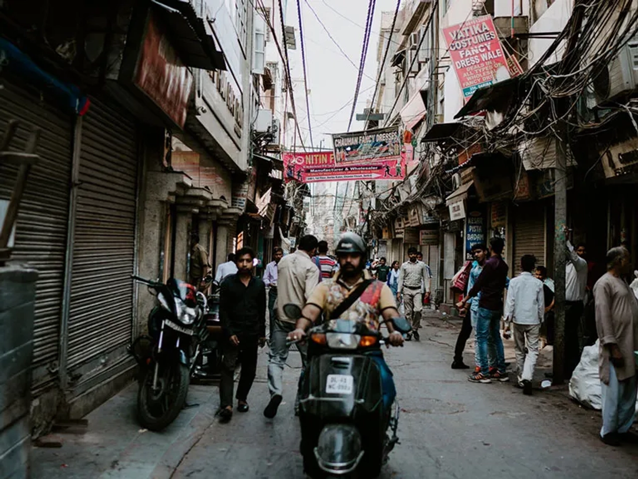 Rua movimentada em Nova Deli, com pessoas e motocicletas entre lojas fechadas.
