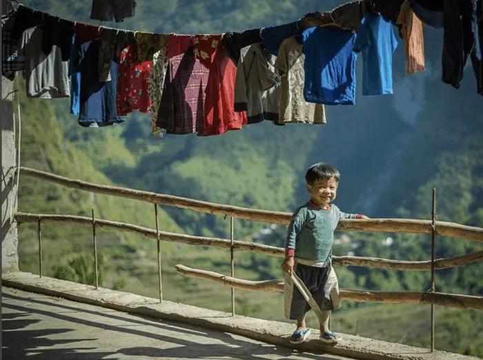 Niño sonriente con un palo, ropa colgada y paisaje montañoso de fondo.