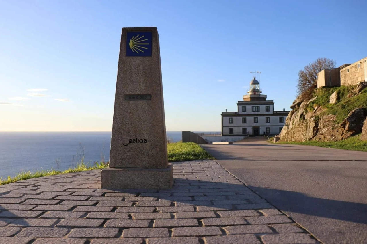Marco do Caminho de Santiago em Galícia, com vista para o mar e farol ao fundo.