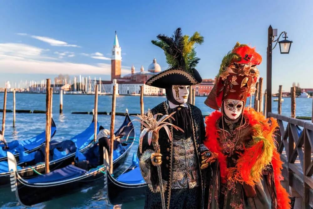 duas pessoas vestidas com trajes de carnaval posam em frente às gondolas e a um dos canais de veneza