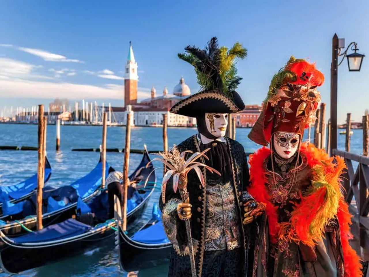 duas pessoas vestidas com trajes de carnaval posam em frente às gondolas e a um dos canais de veneza