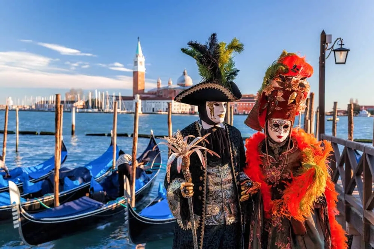 duas pessoas vestidas com trajes de carnaval posam em frente às gondolas e a um dos canais de veneza