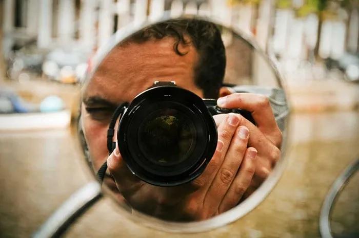 Fotógrafo reflejado en un espejo, sosteniendo una cámara. Fondo urbano desenfocado.