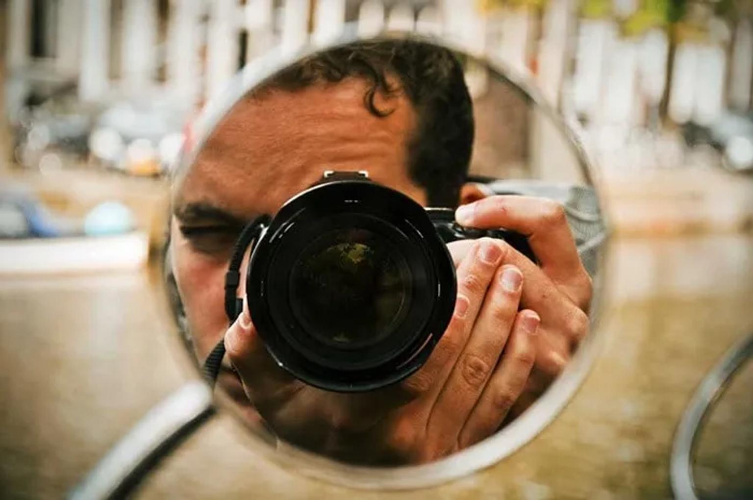 Fotógrafo reflejado en un espejo, sosteniendo una cámara. Fondo urbano desenfocado.