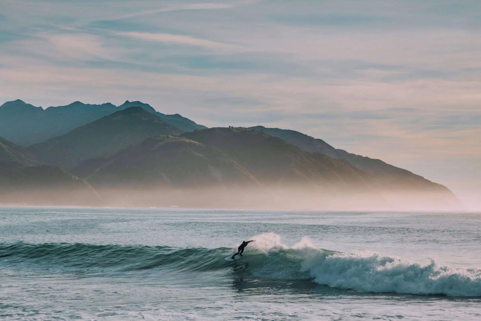 Surfista pegando onda em um mar tranquilo, com montanhas ao fundo.