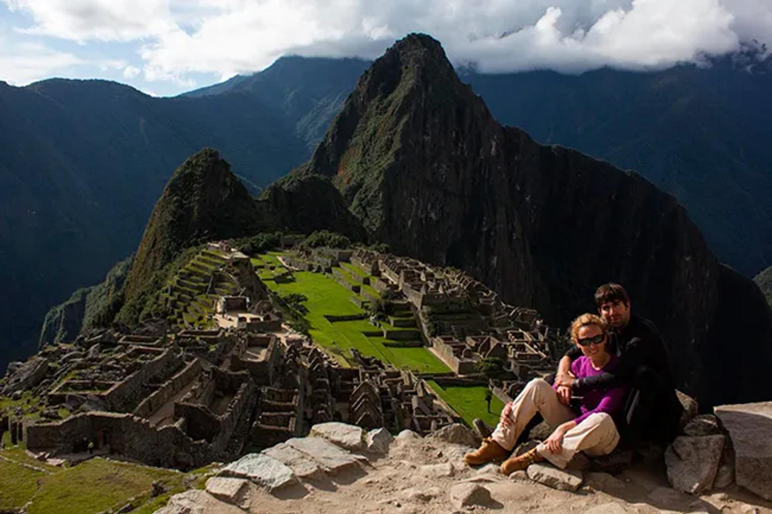 Pareja sentada en Machu Picchu, con montañas y ruinas al fondo.