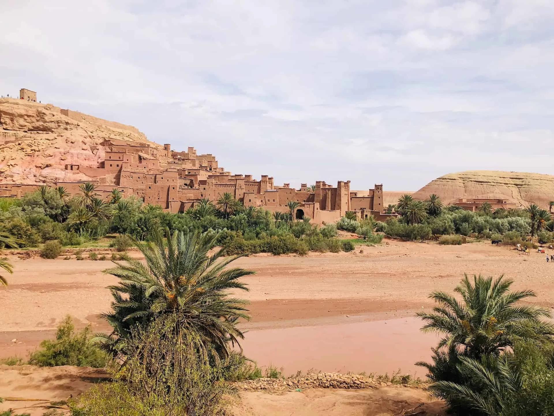 A traditional Moroccan village with clay buildings surrounded by palm trees and mountains.