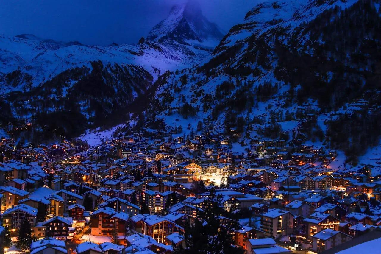 Vista noturna de uma cidade alpina iluminada, cercada por montanhas cobertas de neve.