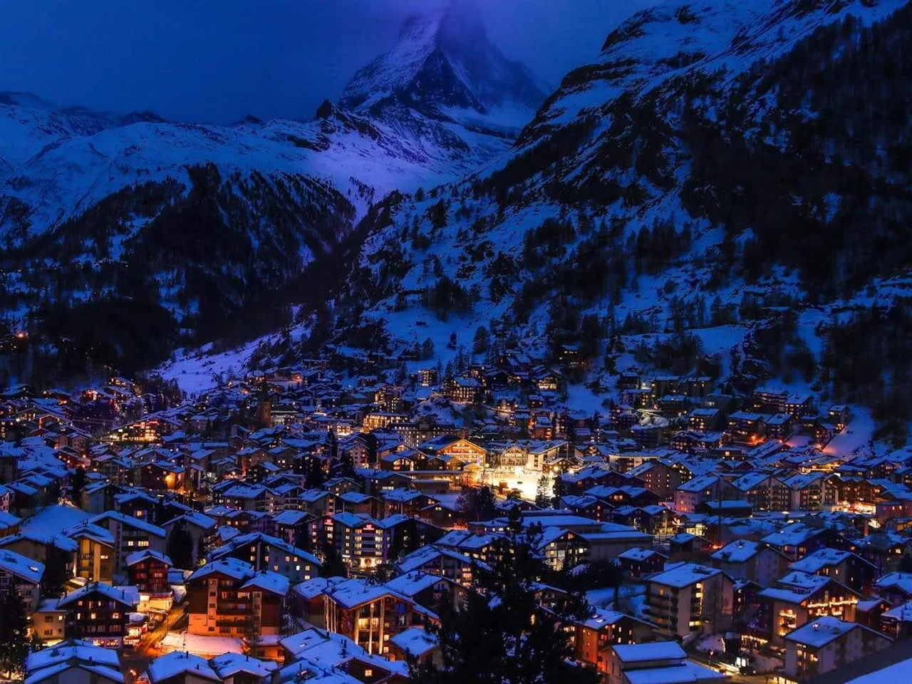 Vista noturna de uma cidade alpina iluminada, cercada por montanhas cobertas de neve.