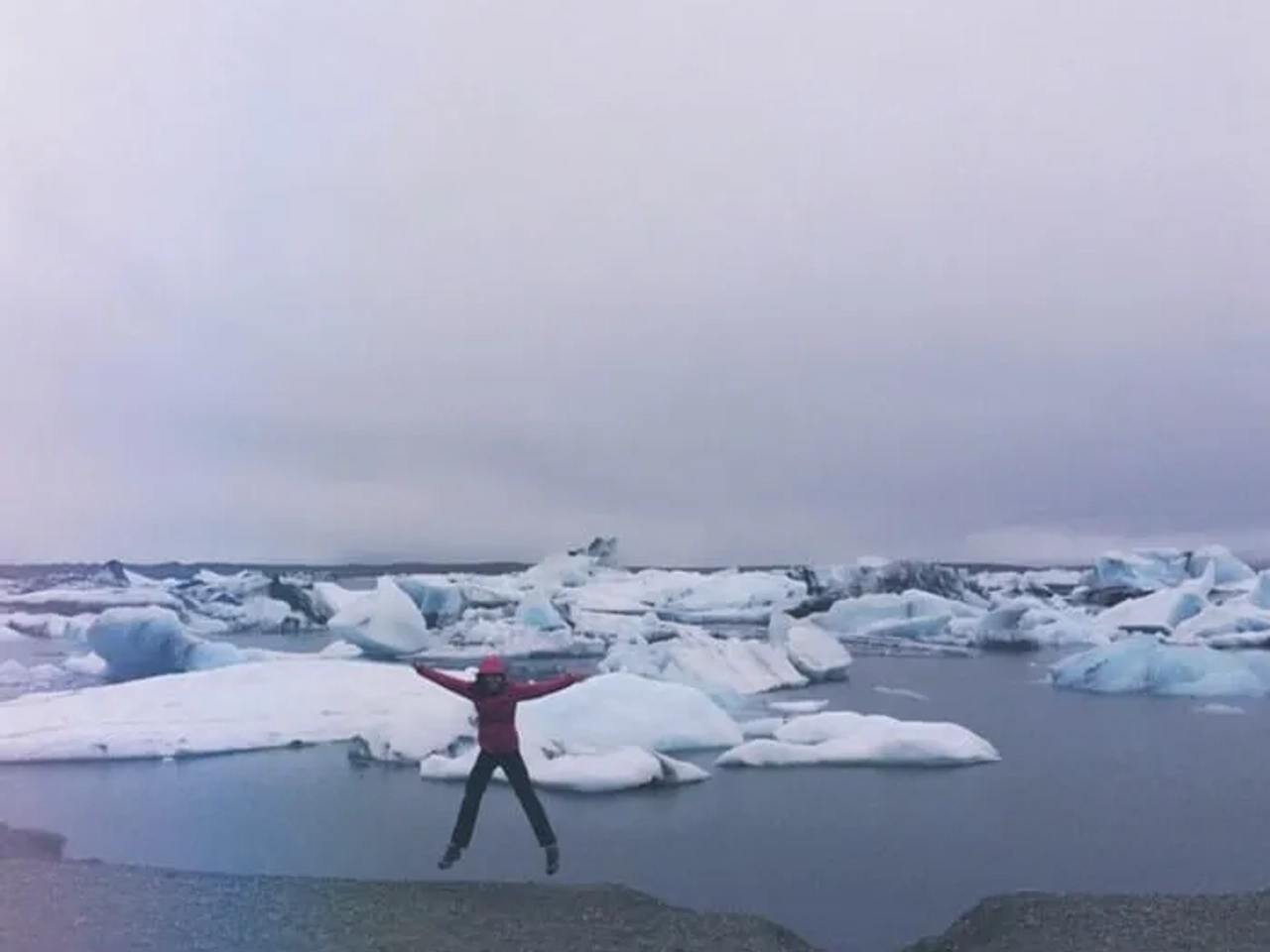 Mujer saltando frente a icebergs en un paisaje helado y nublado.