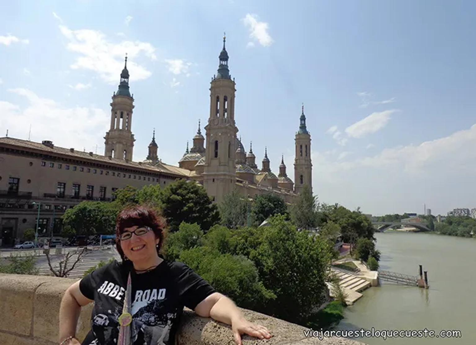 Mujer sonriente frente a un edificio histórico y un río en un día soleado.