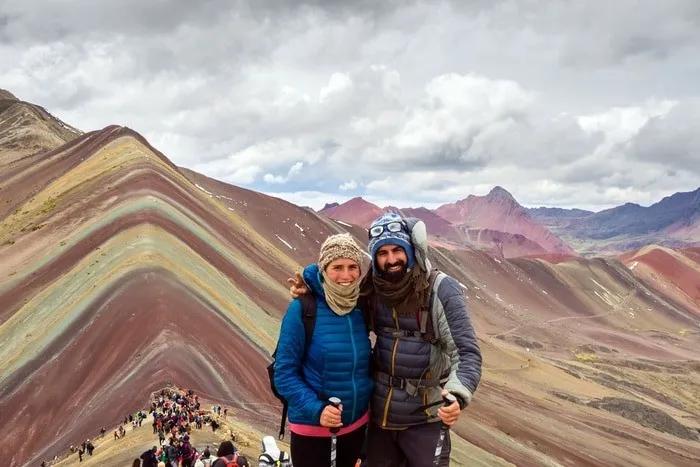 Pareja sonriente en la montaña de colores, rodeada de turistas y paisajes impresionantes.