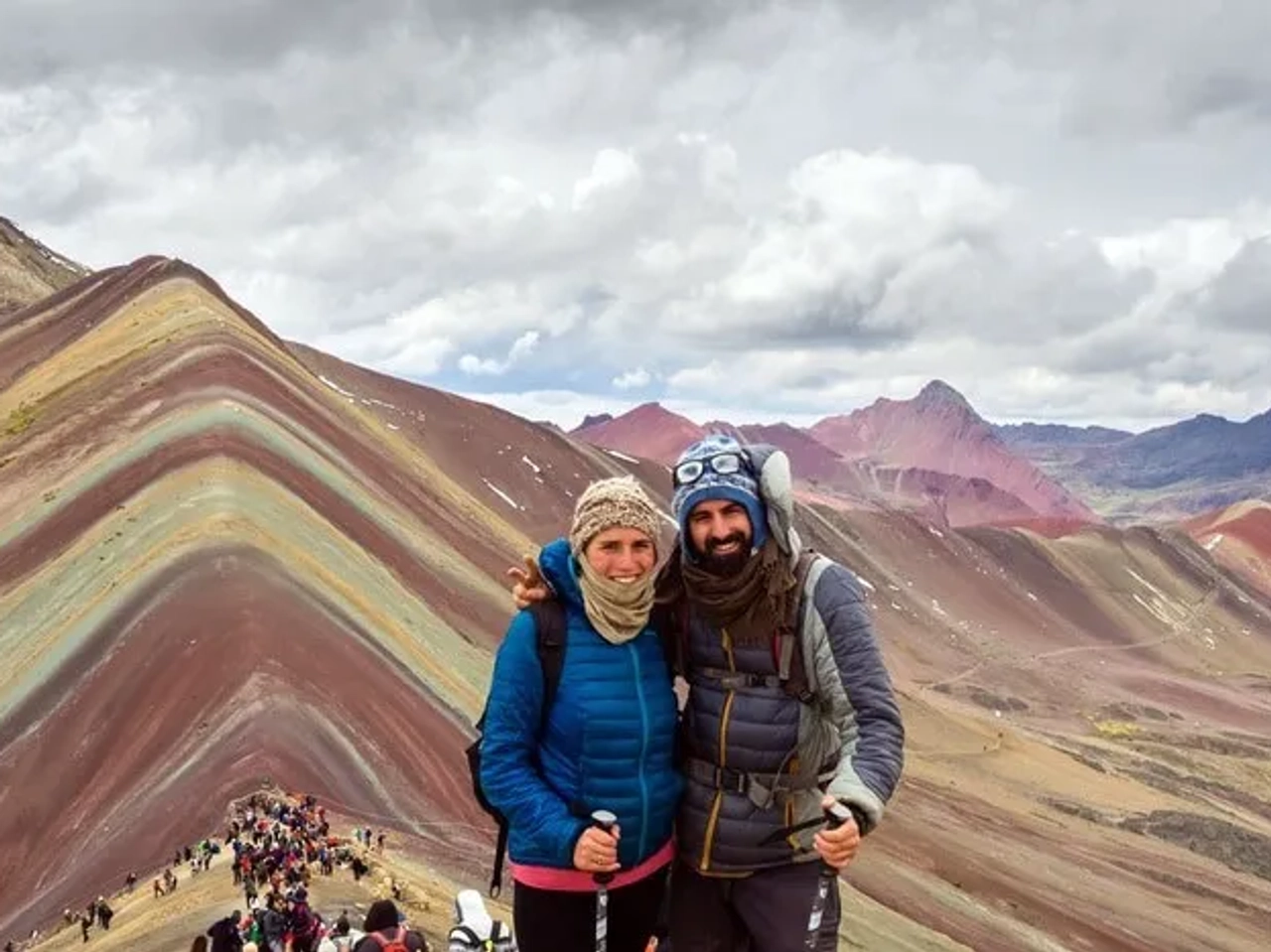 Pareja sonriente en la montaña de colores, rodeada de turistas y paisajes impresionantes.