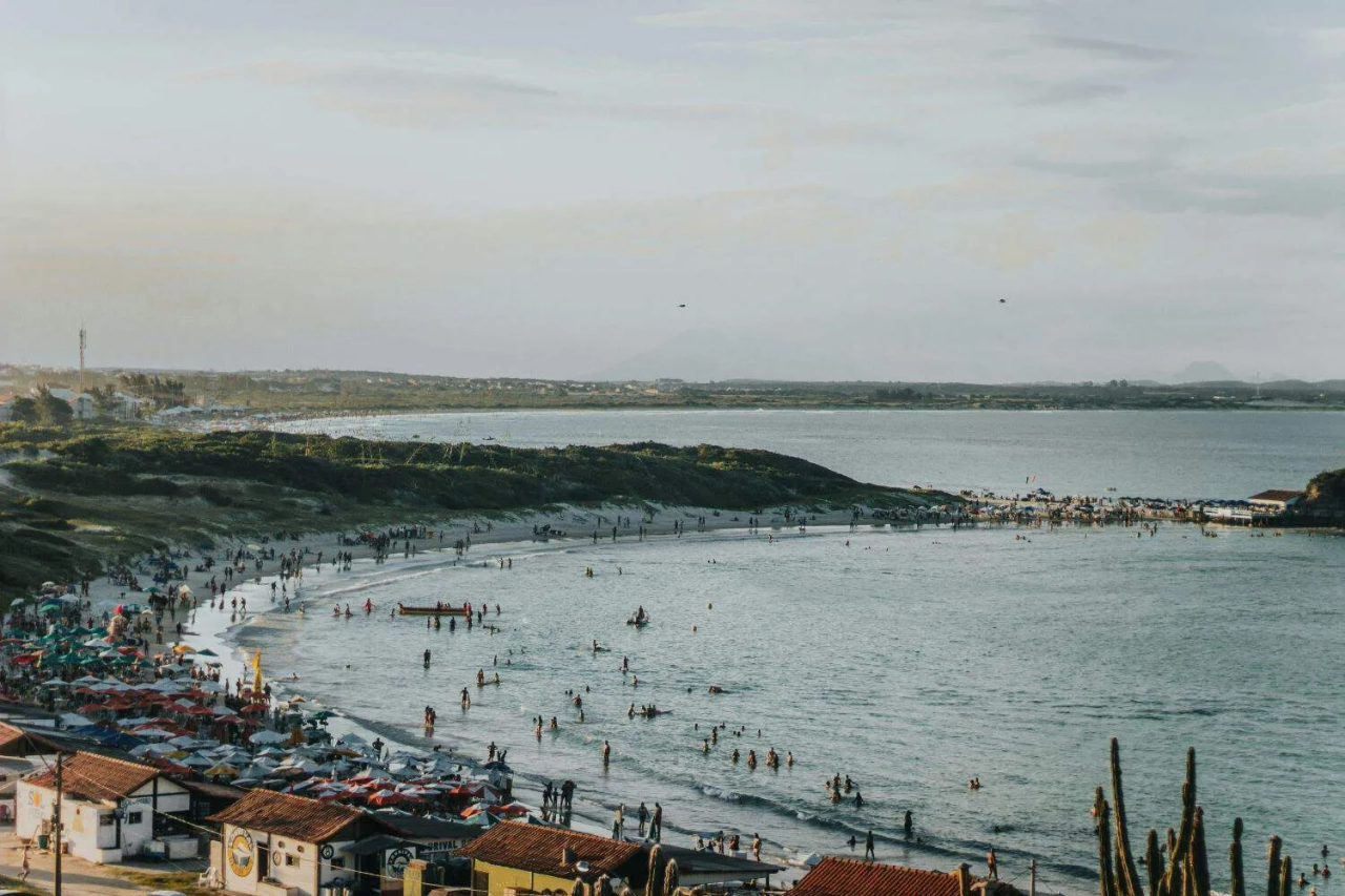 Praia movimentada com pessoas, barracas coloridas e mar calmo ao fundo.