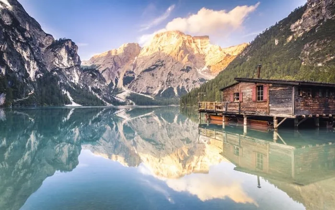 Lago alpino em Itália com montanhas refletidas na água e uma casa de madeira junto à margem.