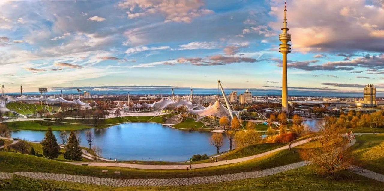 Vista panorâmica do Parque Olímpico de Munique, com lago e torres icônicas.