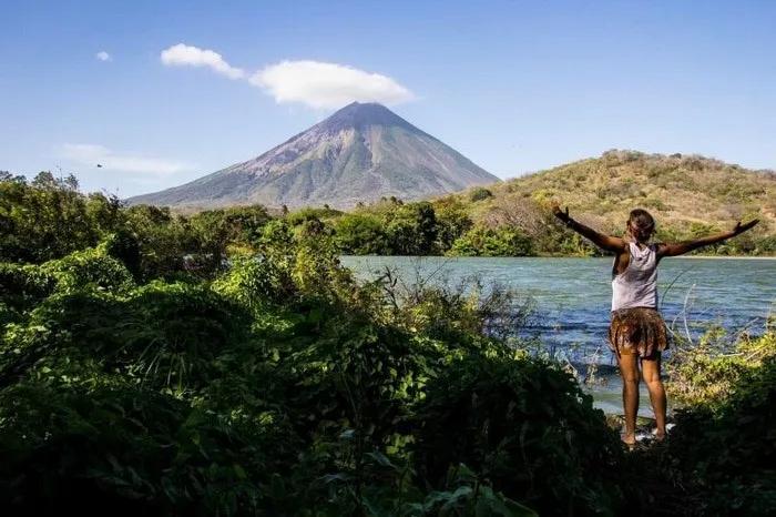 Mujer de espaldas frente a un volcán y un lago rodeado de vegetación.