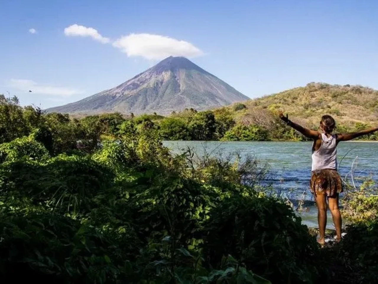 Mujer de espaldas frente a un volcán y un lago rodeado de vegetación.