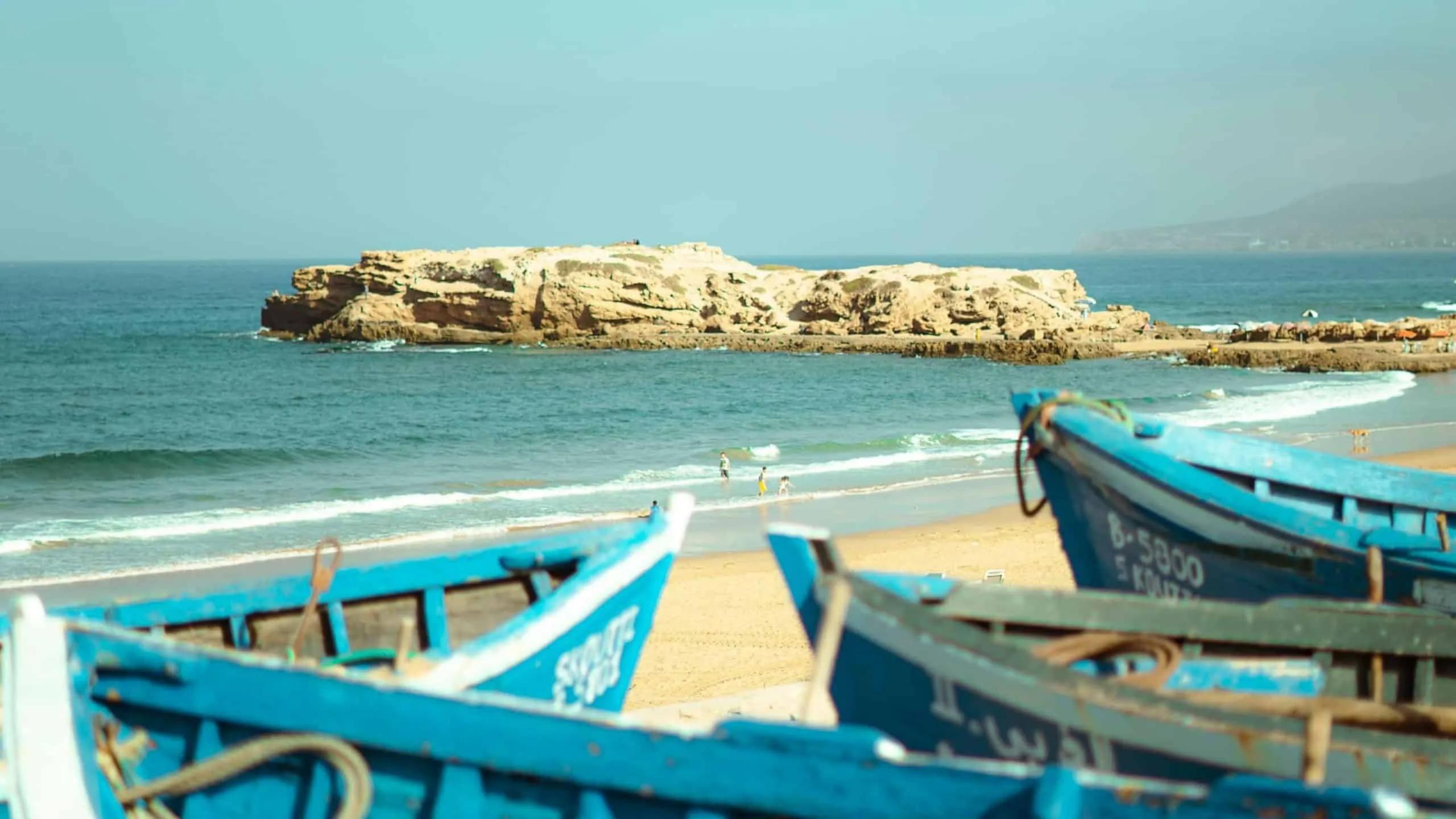 Praia em Marrocos com barcos azuis e rochas ao fundo.